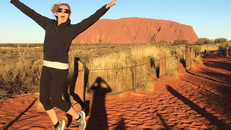 Excited woman jumps on vibrant red dirt track by Uluru, Australia, during a 3-night Rock The Centre Yulara to Yulara tour.