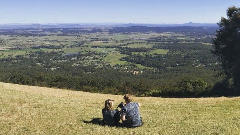 Two people relax on a lush grassy hill overlooking scenic Gold Coast valleys, envisioning a luxurious Ultimate Gourmet Tasting Experience.