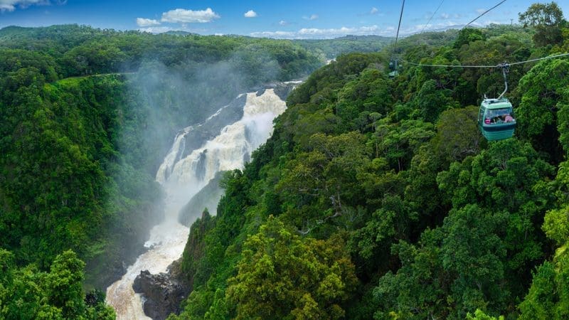 Skyrail cable car travels above vibrant rainforest canopy, overlooking a majestic waterfall under a bright blue sky with fluffy clouds.