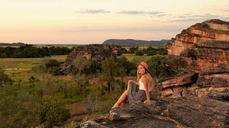 Woman wearing a sunhat admires vibrant Kakadu sunset from a scenic rocky ledge on her 1 Day Wilderness Escape with Crocodile Cruise.