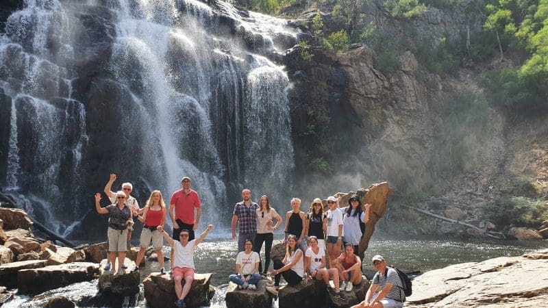 A group enjoys scenic rocks by a majestic Grampians waterfall, framed by lush trees and rugged cliffs on their day tour adventure.