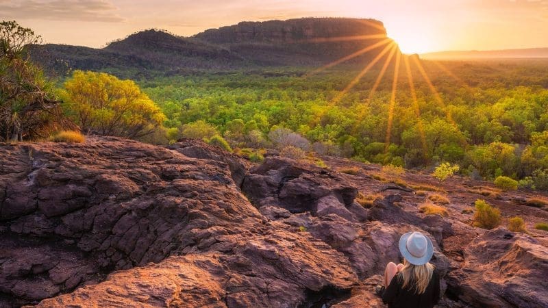 Woman wearing a hat sits on rugged rocks at sunset during a 4-Day Kakadu Katherine Gorge Adventure, enjoying breath-taking views.