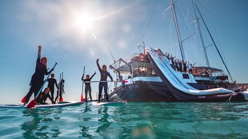 Group in wetsuits paddleboard and celebrate by a luxury yacht with a waterslide on clear blue water under bright sun.