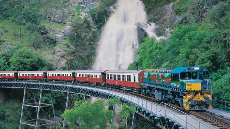 Kuranda Scenic Railway’s colourful Kuranda Deluxe train crosses a bridge near a waterfall and vibrant, lush green rocky hills in Australia.