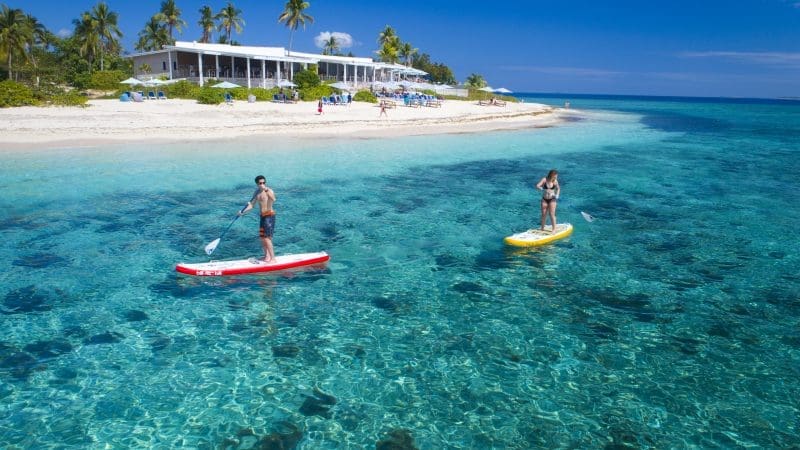 Two paddleboarders glide on crystal-clear turquoise water by a tropical sandy beach with palm trees and a white resort building.