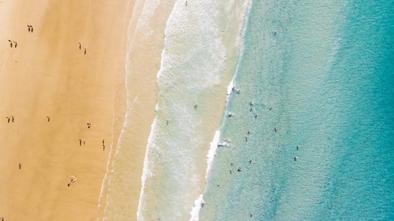 Aerial drone shot of beachgoers relaxing on golden sand, some swimming in crystal clear turquoise sea waves under bright sunny skies.