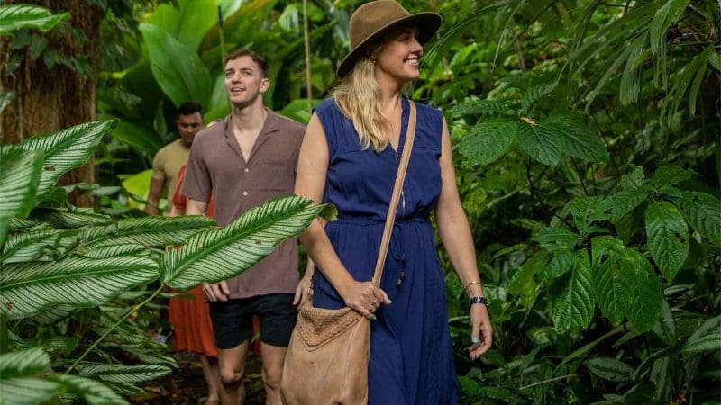 Two people smile as they walk among lush tropical plants on a CSHD Tour, with more following in Cairns City.