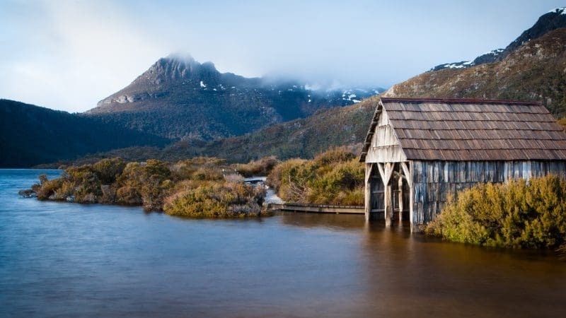 Rustic wooden boathouse on tranquil lake shore with majestic mountain backdrop, serene water reflections, and scenic natural landscape.
