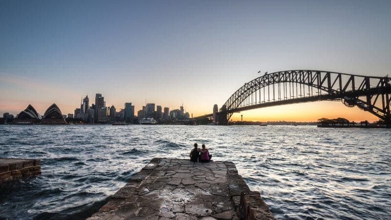 Two people sit on a stone pier at sunset, gazing at Sydney Opera House and Harbour Bridge over sparkling harbour water, iconic Australia.