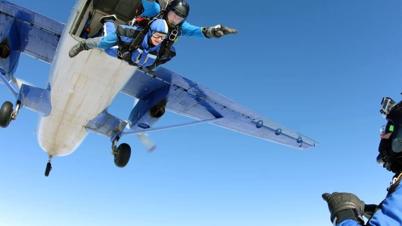 a skydiving instructor and tandem partner jumping out of an airplane with the plane in the background and blue skies
